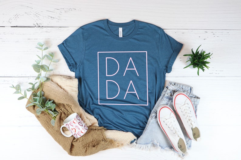 May include: A blue t-shirt with the word "DADA" in a pink square. The shirt is styled with a pair of denim shorts, white sneakers, a mug with heart designs, and a folded brown sweater. The items are arranged on a white wooden surface.