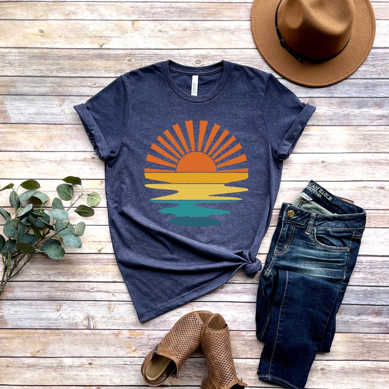 May include: A navy blue t-shirt with a retro sunset graphic featuring a yellow sun with orange rays and a blue, green, and yellow reflection in the water. The t-shirt is laid flat on a wooden surface with a pair of brown sandals, a pair of blue jeans, and a brown fedora hat.