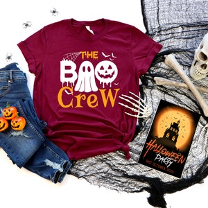 May include: A maroon t-shirt with the text "The Boo Crew" in white and orange. The text includes a ghost and a jack-o-lantern. The shirt is laid out on a white surface with a pair of blue jeans, a skull, and a Halloween party invitation.