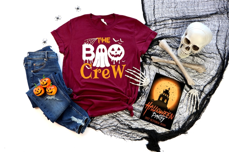 May include: A maroon t-shirt with the text "The Boo Crew" in white and orange. The text includes a ghost and a jack-o-lantern. The shirt is laid out on a white surface with a pair of blue jeans, a skull, and a Halloween party invitation.