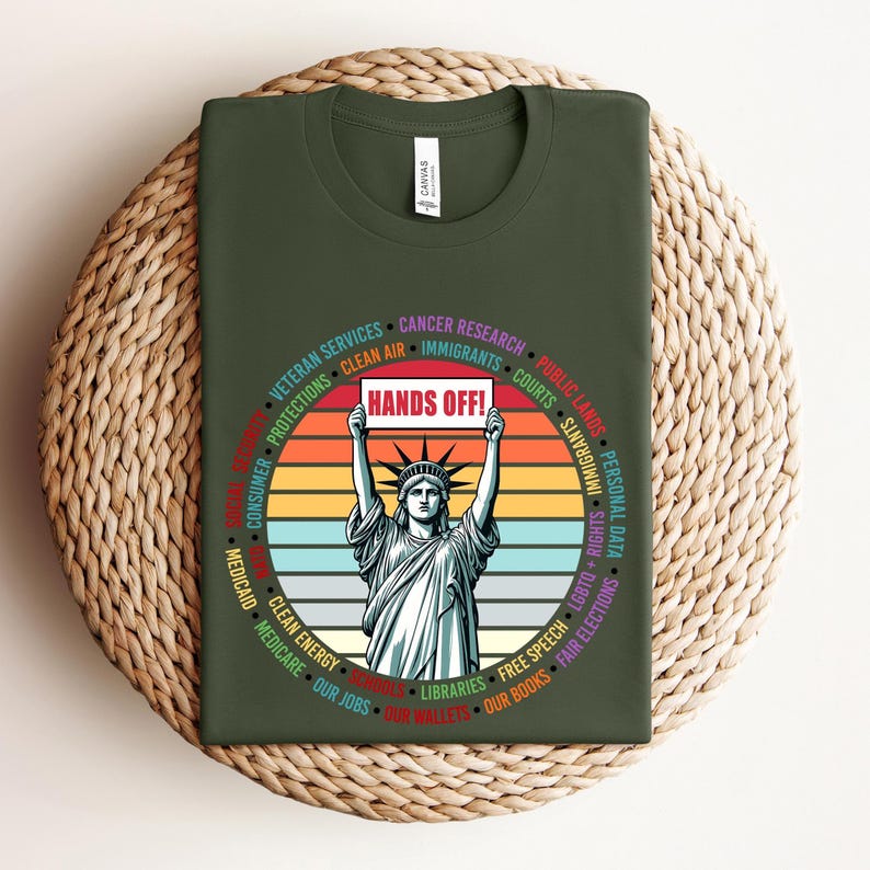 May include: Green t-shirt with a vintage-style graphic of the Statue of Liberty holding a sign that says "Hands Off!"  The graphic is surrounded by a circle of text listing various things the wearer believes should be protected, such as "Clean Air", "Medicare", "Our Jobs", "Libraries", and "Free Speech".