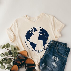 Peut inclure: Un t-shirt cr&egrave;me avec un motif de globe terrestre bleu fonc&eacute; et le mot "travel" &eacute;crit en cursive en dessous. Le t-shirt est associ&eacute; &agrave; un short en jean et des sandales.