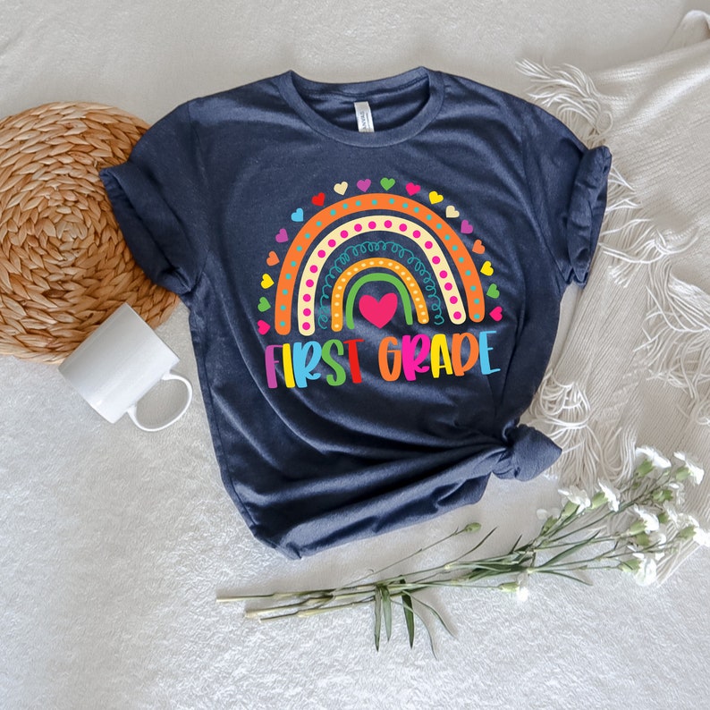 Peut inclure: Un t-shirt bleu marine avec un motif arc-en-ciel et le texte "First Grade" en lettres color&eacute;es. L'arc-en-ciel a un c&oelig;ur au centre et est entour&eacute; de petits c&oelig;urs.