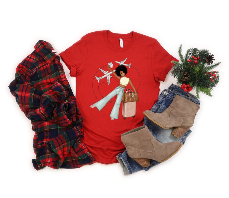 Puede incluir: Camiseta roja con un gr&aacute;fico de una mujer con afro sosteniendo una maleta y una tarjeta de embarque, con aviones volando por encima. La mujer lleva pantalones vaqueros azules y una camiseta blanca.