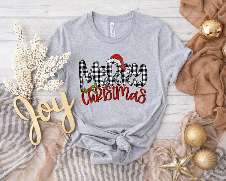 May include: Gray t-shirt with a black and white buffalo plaid design and the text "Merry Christmas" in red with a Santa hat on top of the "M".