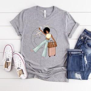 Puede incluir: Una camiseta gris con un gr&aacute;fico de una mujer con cabello casta&ntilde;o y una maleta, sosteniendo un pasaporte y una tarjeta de embarque, con aviones volando a su alrededor. La mujer lleva una blusa amarilla, pantalones vaqueros azules y sandalias marrones.