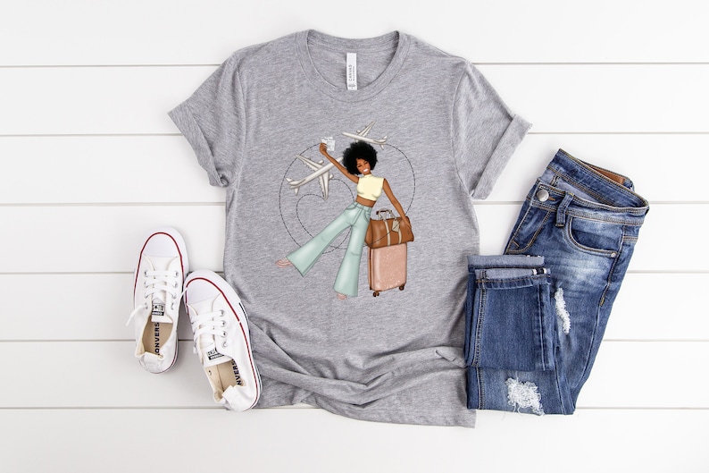 Puede incluir: Una camiseta gris con un gr&aacute;fico de una mujer con cabello casta&ntilde;o y una maleta, sosteniendo un pasaporte y una tarjeta de embarque, con aviones volando a su alrededor. La mujer lleva una blusa amarilla, pantalones vaqueros azules y sandalias marrones.