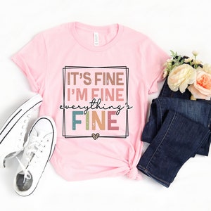 May include: Pink t-shirt with the text "It's fine I'm fine everything's fine" in a colorful, distressed font.