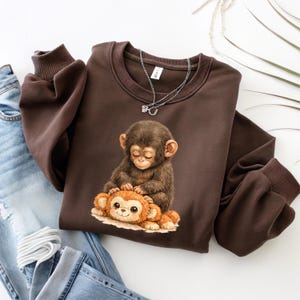 May include: A brown sweatshirt featuring a graphic of a baby monkey with closed eyes, gently touching a smaller, orange monkey. The sweatshirt has a crew neck and long sleeves. A silver necklace with a small pendant is draped on the sweatshirt.
