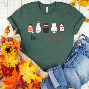May include: A green t-shirt with a festive design featuring five cats wearing Christmas hats and ornaments. The text "Meowy Christmas" is printed below the cats.