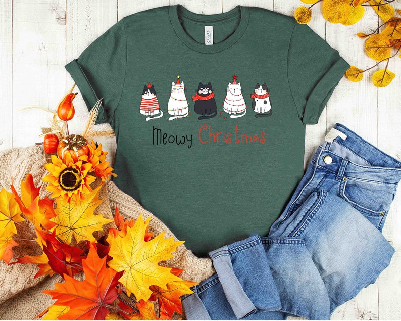 May include: A green t-shirt with a festive design featuring five cats wearing Christmas hats and ornaments. The text "Meowy Christmas" is printed below the cats.