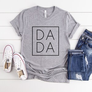 May include: A gray t-shirt with the word "DADA" in a black square, paired with blue jeans and white sneakers. The shirt is laid on a white wooden surface, creating a casual, everyday look.