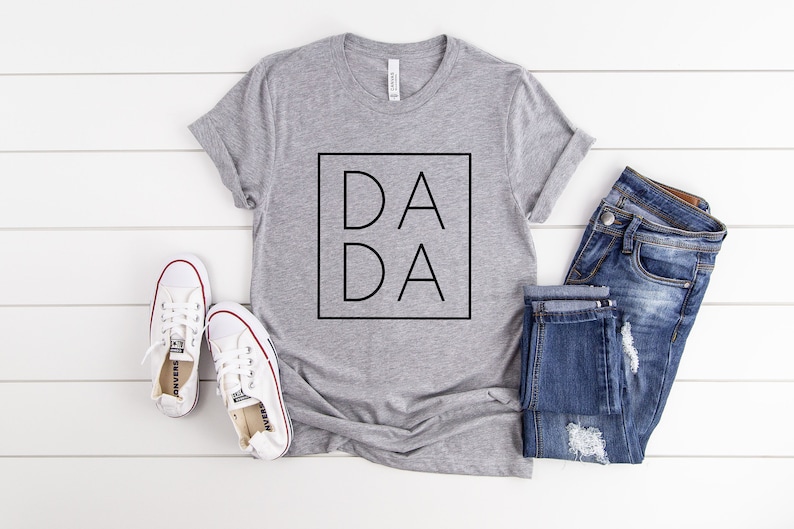 May include: A gray t-shirt with the word "DADA" in a black square, paired with blue jeans and white sneakers. The shirt is laid on a white wooden surface, creating a casual, everyday look.