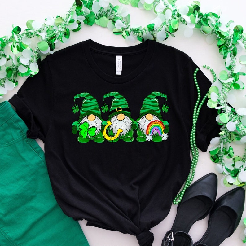 Peut inclure: T-shirt noir avec un motif de trois lutins portant des chapeaux verts et tenant un tr&egrave;fle, un fer &agrave; cheval et un arc-en-ciel. Les lutins sont sur un fond noir.