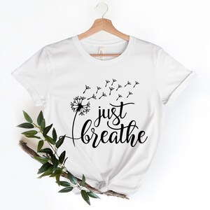Op de afbeelding: Wit T-shirt met een zwarte paardenbloem afbeelding en de tekst "just breathe" erop gedrukt.