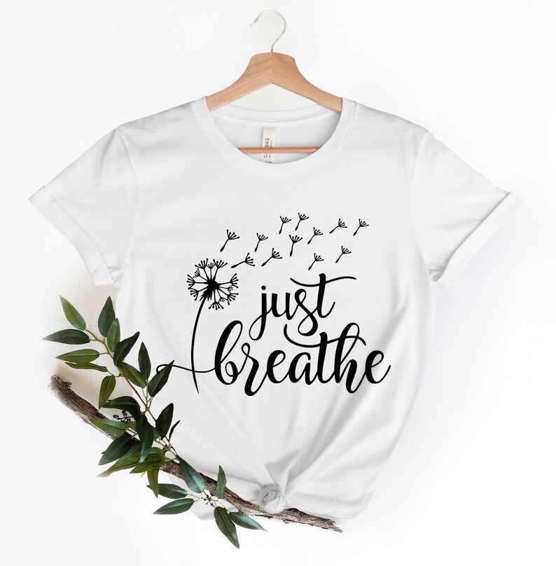 Op de afbeelding: Wit T-shirt met een zwarte paardenbloem afbeelding en de tekst "just breathe" erop gedrukt.