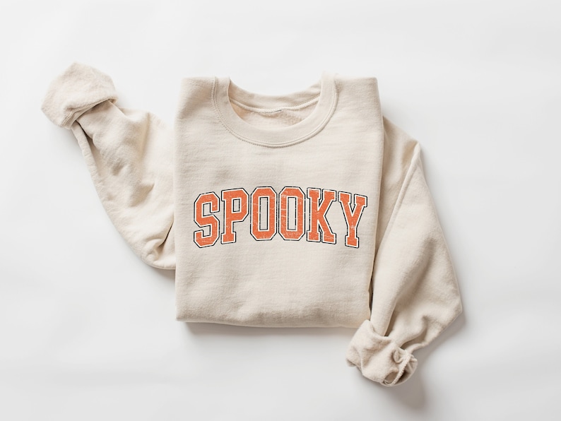 May include: A beige sweatshirt with the word "SPOOKY" printed in orange with black outlines. The sweatshirt is folded and laid flat on a white surface.