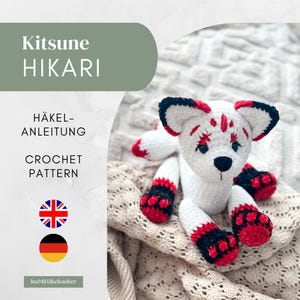 Könnte beinhalten: Eine Häkelanleitung für ein Kitsune Hikari Fuchs-Spielzeug. Der Text lautet Kitsune HIKARI, HÄKEL-ANLEITUNG und CROCHET PATTERN. Das Spielzeug ist weiß mit schwarzen und roten Akzenten an Ohren, Pfoten und Schwanz sowie einer schwarzen Nase.