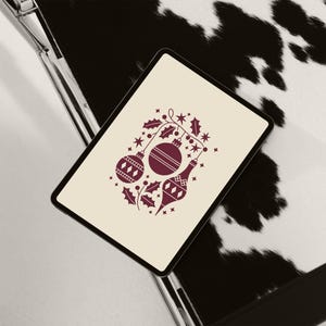 May include: A digital tablet displays a festive Christmas ornament design. The design features burgundy-colored ornaments, holly, and stars on a cream background. The tablet rests on a black and white cowhide pattern.