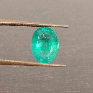May include: A vibrant, oval-cut emerald gemstone held by tweezers. The gem displays a rich, saturated green color with visible facets. The background is a dark, neutral tone, highlighting the gemstone's brilliance.