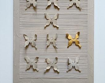 May include: A beige rectangular canvas with a textured surface and a raised border. Nine butterfly sculptures are arranged in a grid pattern, with one gold-colored butterfly. The butterflies have detailed wing patterns.