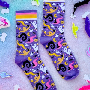May include: A pair of purple socks with a Halloween-themed pattern. The socks feature ghosts, witch hats, candy corn, and swirling worm designs in white, yellow, black, and pink. The top band is striped with yellow, pink, and white.
