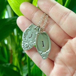 Könnte beinhalten: Eine silberne Halskette mit zwei Anhängern. Ein Anhänger ist löffelförmig und hat den Buchstaben "J" eingraviert. Der andere Anhänger ist oval und hat die Zahl "1" eingraviert.