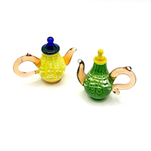 May include: Two miniature glass teapots. One is yellow with a blue and green lid, and the other is green with a yellow lid. Both have clear handles and spouts. The teapots are decorative and likely for display.