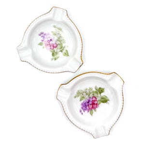 May include: Two white porcelain ashtrays with a floral design. Each ashtray has a gold trim and a painted bouquet of purple, pink, and white flowers. The ashtrays are triangular with three rests for cigarettes.