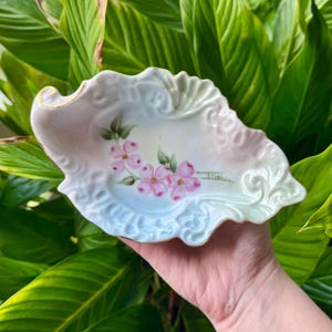 May include: A hand-painted, porcelain dish with a scalloped edge and gold trim. The dish features a pastel color gradient and a floral design of pink blossoms and green leaves. The artist's signature, "ETHEL", is visible.