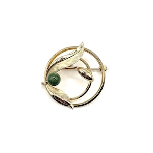 May include: A gold-toned brooch with a circular design featuring stylized leaves and a green gemstone. The brooch has a pin closure on the back. The leaves and gemstone are the focal points of the design.
