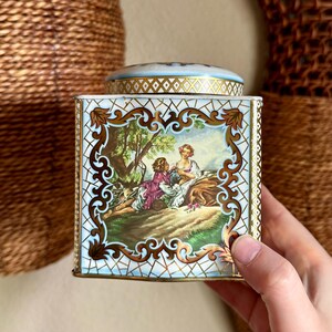 Puede incluir: Una lata cuadrada vintage con tapa. Presenta una pintura de dos figuras en una escena de jardín, enmarcada por filigranas doradas. La tapa es azul claro con un patrón geométrico dorado. La lata es sostenida por una mano.
