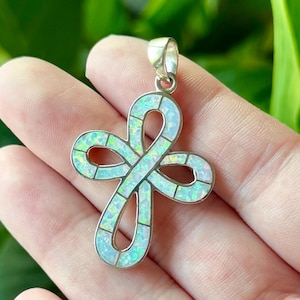 Puede incluir: Colgante de plata con forma de cruz con incrustaciones de ópalo. El ópalo muestra un brillo iridiscente, con toques de azul, verde y amarillo. El colgante tiene un lazo en la parte superior para una cadena. Mide unos 3 cm.