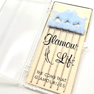 May include: A vintage hair comb in a clear plastic case. The comb has silver metal teeth and a light blue decorative top with a camel design. The text "Glamour Lift" and "The comb that glamourizes" are printed on the card.