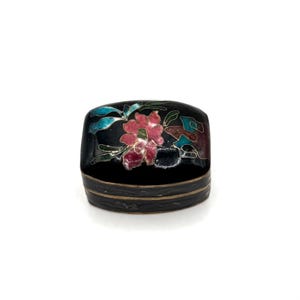 May include: A small, black, rectangular trinket box with a floral design. The box features a pink flower with green leaves and blue accents. The box has a gold-colored band around the base.