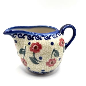 May include: Creamer with a blue handle and spout, decorated with red flowers and green leaves. The rim and handle are blue, and the body has a dotted pattern. A charming addition to any kitchen.