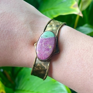 May include: A brass cuff bracelet with a large, oval gemstone. The gemstone is split, with a light green top and a pinkish-purple bottom. The brass has a hammered texture. The bracelet is worn on a wrist.