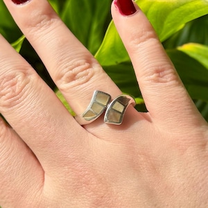 Könnte beinhalten: Ein silberner Ring mit zwei gebogenen, rechteckigen Abschnitten. Jeder Abschnitt hat eine helle, mosaikartige Einlage. Der Ring befindet sich an einem Finger, vor einem grünen Hintergrund.