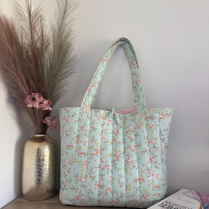 May include: A light blue quilted tote bag adorned with a floral pattern in pink, orange, and yellow. The bag features matching handles and a pink interior. A gold vase with decorative feathers and a book are in the background.