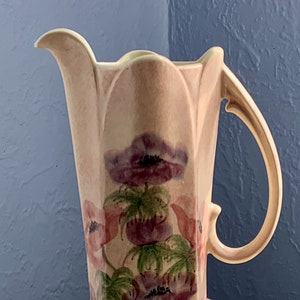 Peut inclure: Un grand pichet en céramique de couleur crème avec un motif floral. Le pichet a une large ouverture et une anse. Le motif floral présente des fleurs roses et violettes avec des feuilles vertes.