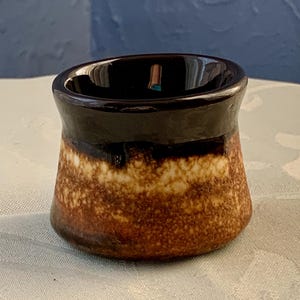 May include: A small, brown and white ceramic egg cup with a dark brown glaze on the top rim.