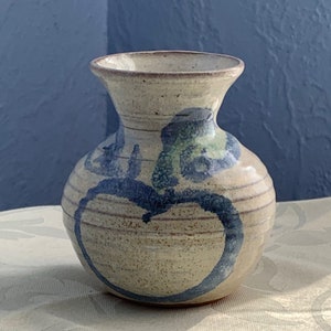May include: A small, hand-painted ceramic vase with a light brown glaze and blue and green abstract designs. The vase has a rounded body and a narrow neck.