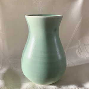 May include: A light green ceramic vase with a rounded bottom and a flared opening. The vase has a subtle ombre effect, transitioning from a lighter shade of green at the top to a darker shade at the bottom.