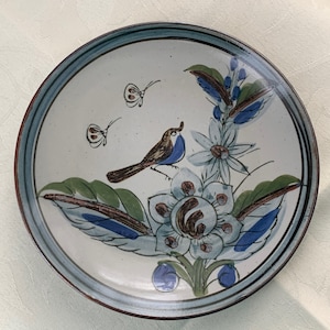 May include: A white ceramic plate with a blue and green floral design and a small blue bird. The plate has a light blue rim.