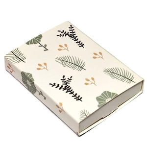Op de afbeelding: Een rechthoekige geschenkdoos met een crèmekleurige achtergrond en een herhalend patroon van groene, zwarte en gouden botanische illustraties. De doos is gesloten en heeft een eenvoudig ontwerp.