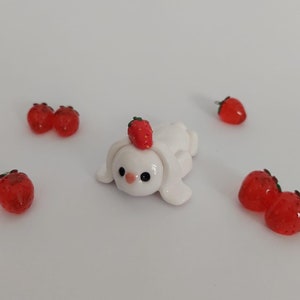 Peut inclure: Une figurine de lapin en céramique blanche avec une fraise rouge sur la tête. Le lapin est couché sur le ventre, les oreilles dressées. Plusieurs fraises rouges sont éparpillées autour du lapin.