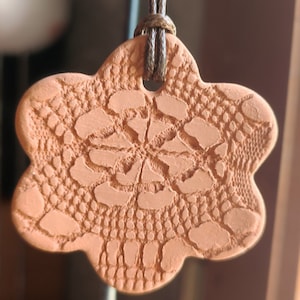 Peut inclure: Un pendentif en terre cuite avec un motif en dentelle détaillé suspendu à un cordon brun.