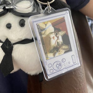 May include: A clear plastic keychain featuring a photo of a cat wearing glasses. The keychain is attached to a plush toy with a black tie and a pair of clear plastic glasses. The photo has the date 2/07/09 18:09.