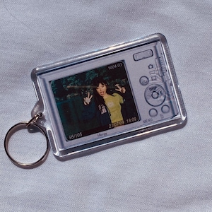 May include: Clear acrylic keychain featuring a photograph of two people making peace signs. The keychain has a gold-colored ring and a design of a camera. The photo has the date 21/07/09 and the time 18:09.