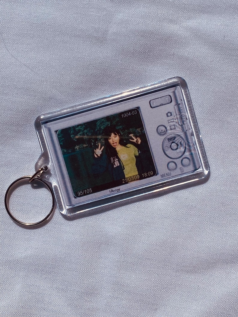 May include: Clear acrylic keychain featuring a photograph of two people making peace signs. The keychain has a gold-colored ring and a design of a camera. The photo has the date 21/07/09 and the time 18:09.